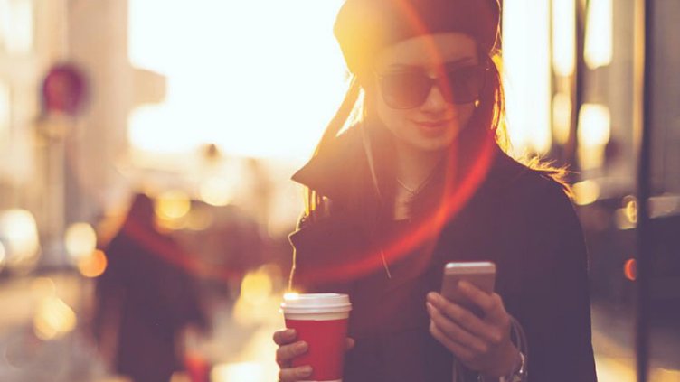 Young woman looking at her phone screen with a cup of coffee in the other hand