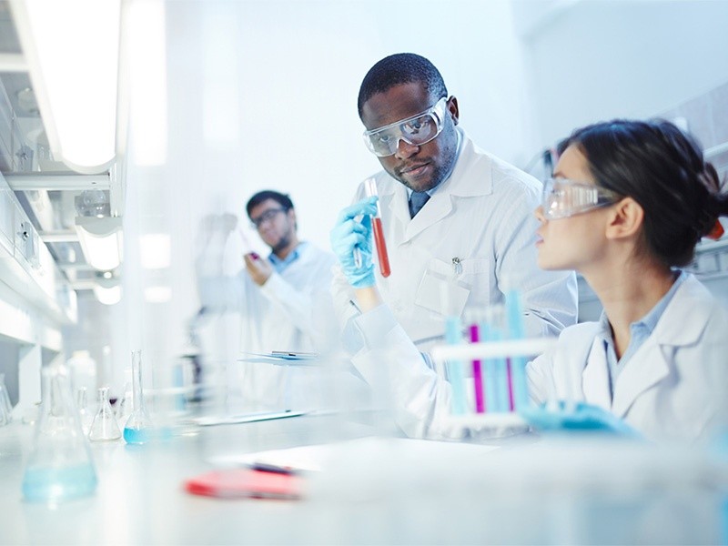 Female Asian laboratory scientist in lab coat and safety goggles showing test tube with red liquid to curious African-American colleague in laboratory. Latin-American scientist in background.