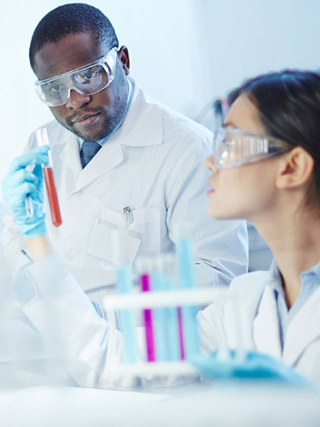 Female Asian laboratory scientist in lab coat and safety goggles showing test tube with red liquid to curious African-American colleague in laboratory. Latin-American scientist in background.