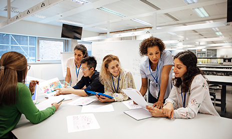 Teacher with a group of university students, in a laboratory classroom. The instructor is considering one of the students work, the mood is light hearted and positive. Other classmates are discussing things with each other. This is a realistic teaching scenario, with candid expressions. This is a multi-ethnic group of women. In the background there is a white board with mathematical formula written on it. All ladies are wearing id tags.