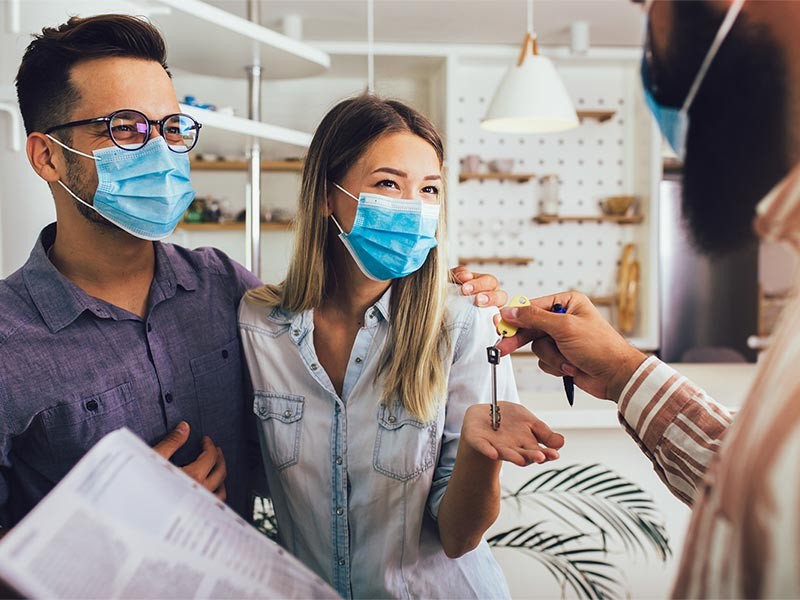 Young happy married couple in medical masks standing in the new house with male real-estate agent. Pandemic concept.