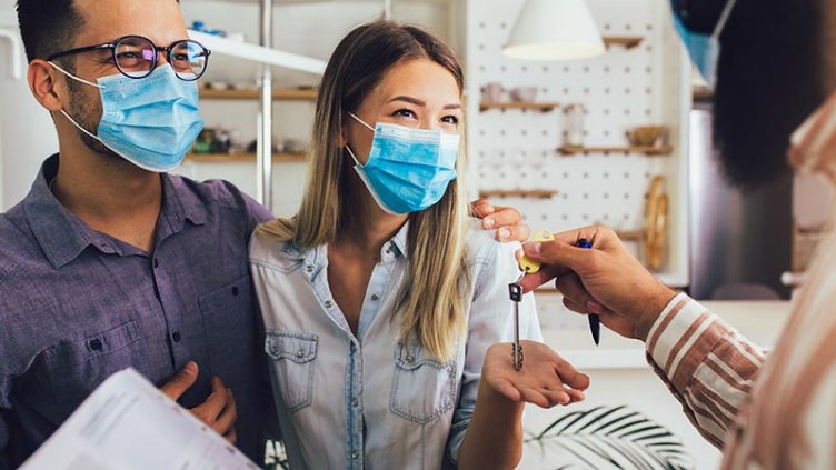 Young happy married couple in medical masks standing in the new house with male real-estate agent. Pandemic concept.