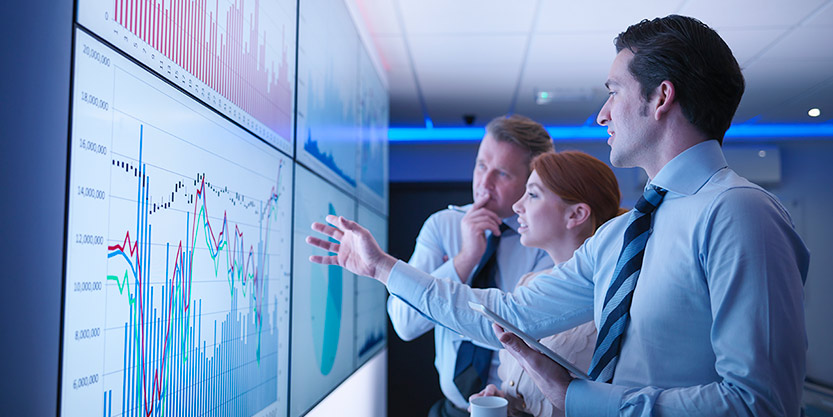 Three business people discuss graphs on screen in meeting room