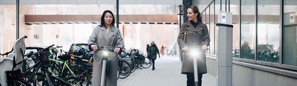 Two business women riding electric scooters on sidewalk in business district