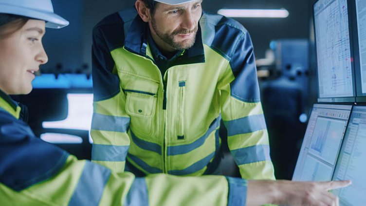 Male and female facilities managers in PPE analyzing smart building data on a bank of computer monitors